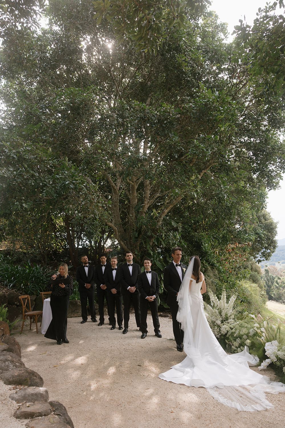 Bride in a long-veiled white gown faces a row of groomsmen in black tuxedos beneath a large tree at an outdoor garden wedding ceremony.
