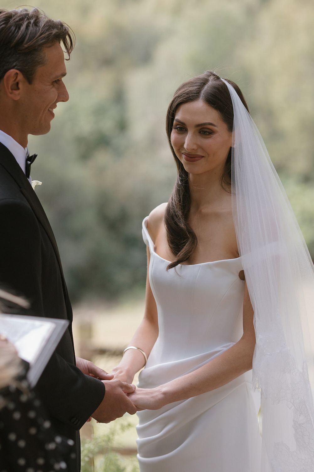 Bride in an elegant white satin gown and lace-trimmed veil holds hands with her groom during an outdoor wedding ceremony.