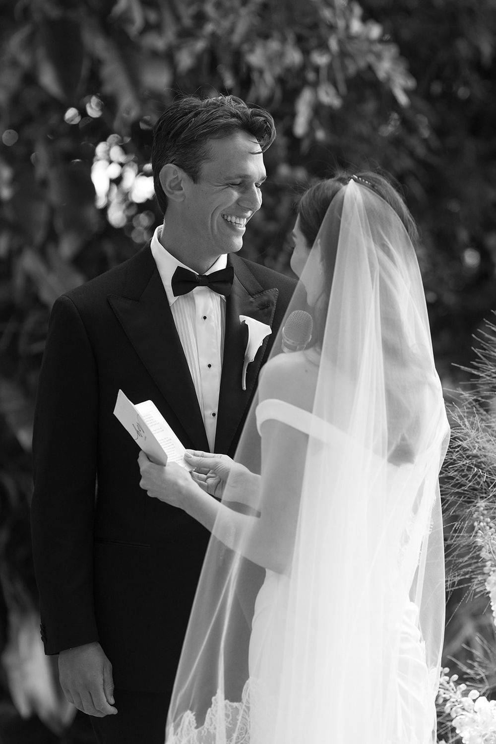 Smiling groom in a black tuxedo and bow tie reads a handwritten note to his bride in a long lace-trimmed veil during an outdoor wedding.