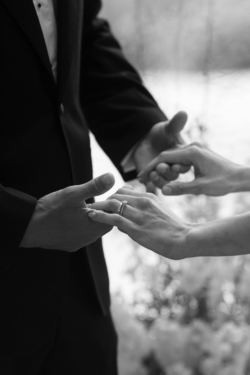 Black and white close-up of a groom in a dark suit holding the bride's hand during a wedding ring exchange ceremony.