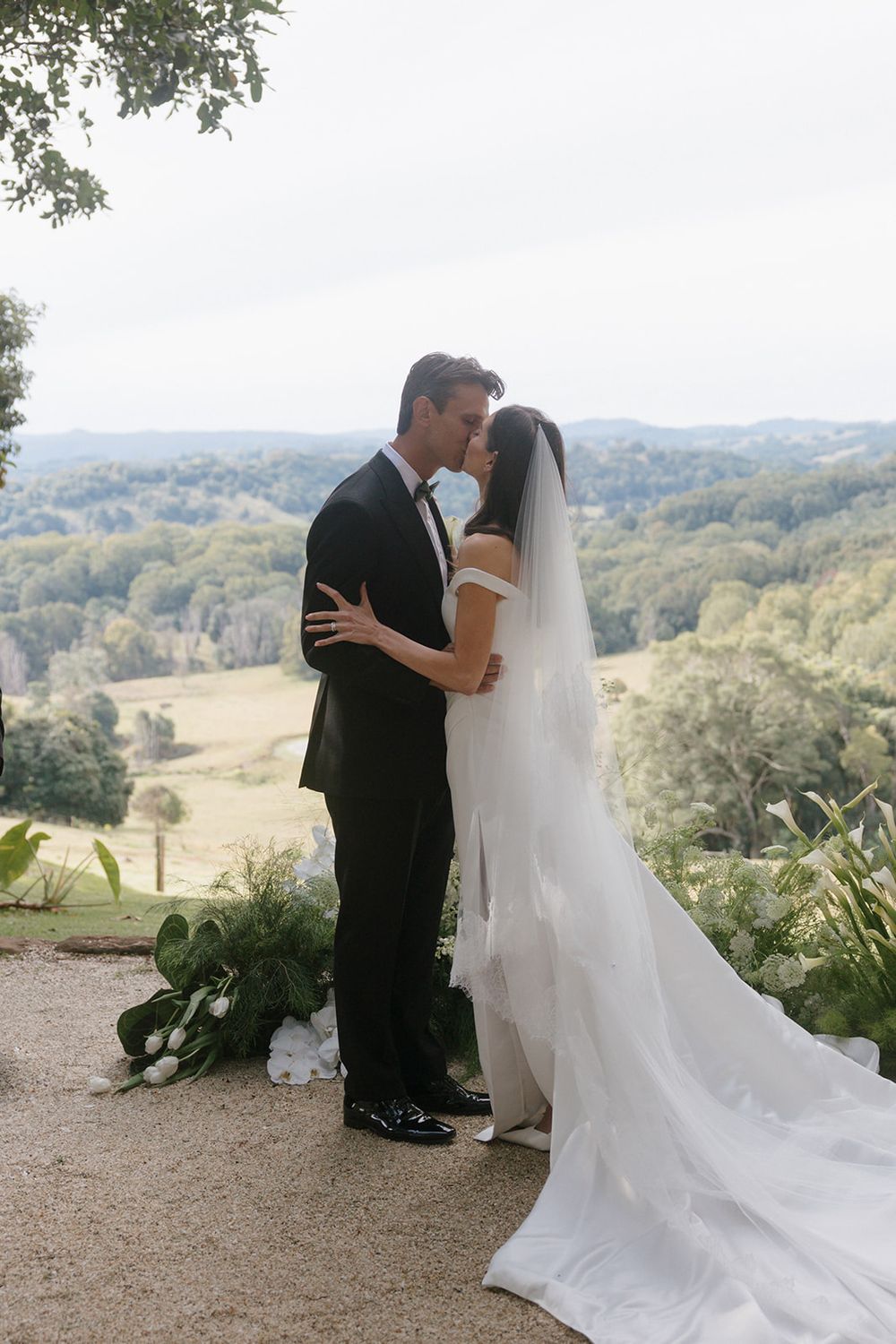 Bride in an off-shoulder gown with a long veil kisses her groom in a black suit on a scenic hilltop overlooking rolling green countryside.