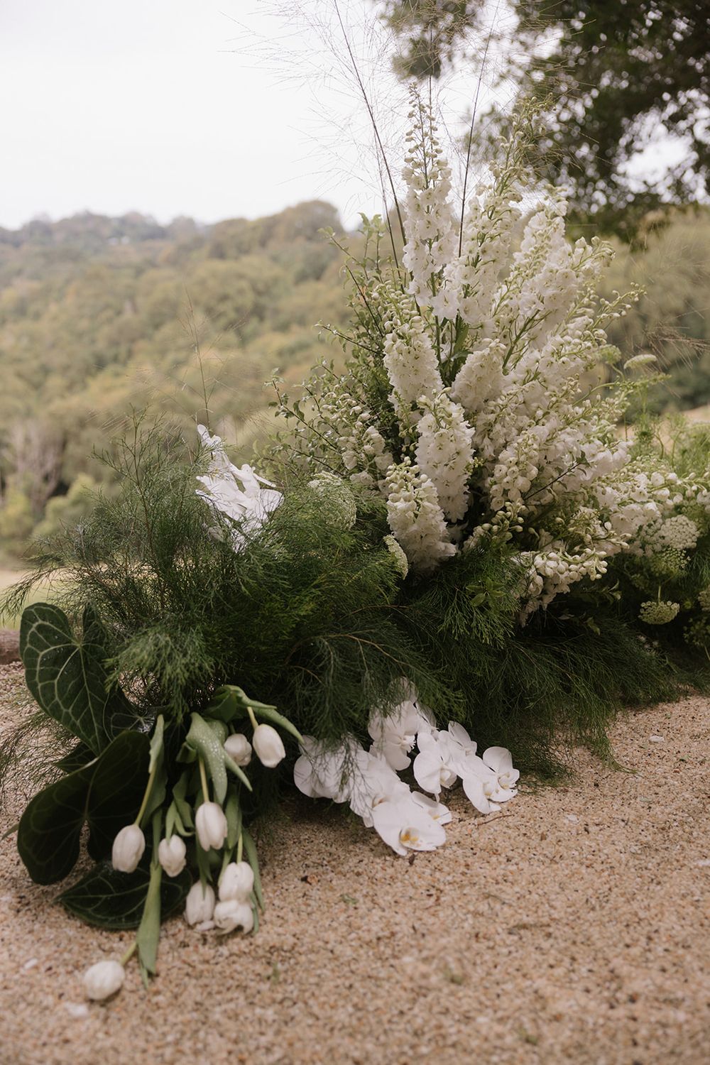 White tulips, orchids and delphinium with lush greenery form a wedding floral arrangement on sand, with forested hills behind.
