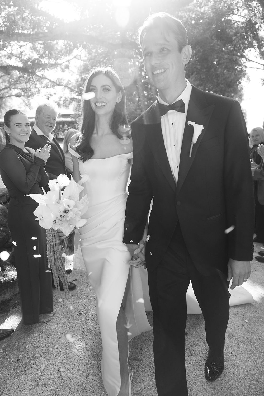 Black and white wedding photo of a smiling bride in a fitted gown and groom in a tuxedo walking hand in hand as guests applaud.