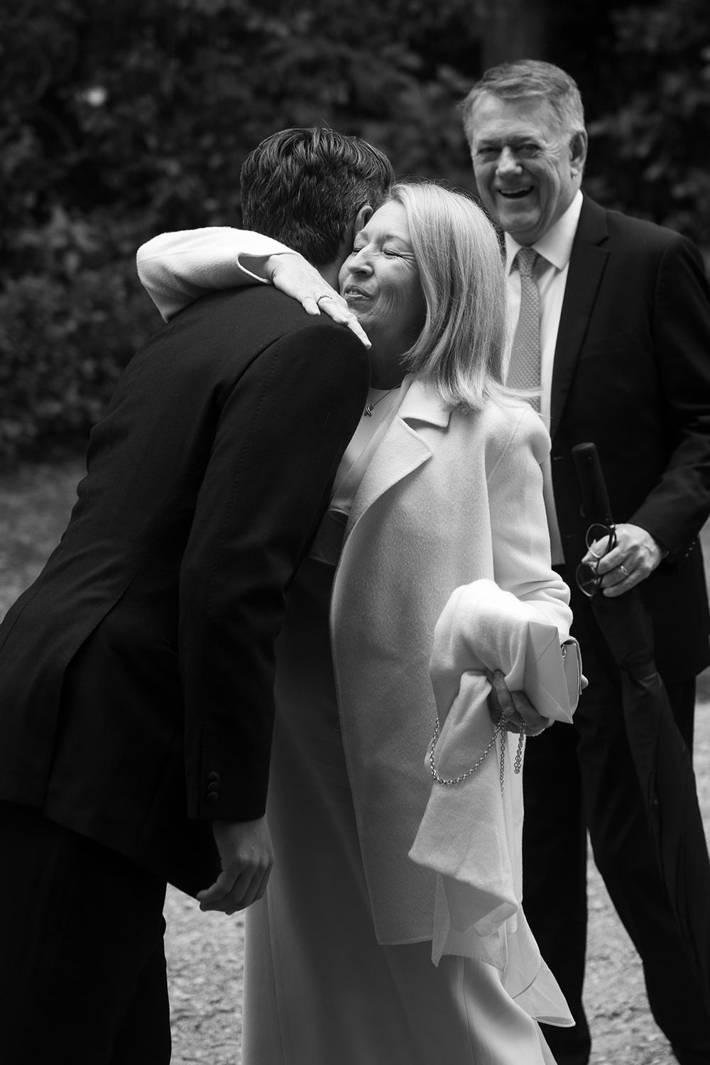 Black and white photo of a blonde woman in a light coat embracing a man in a dark suit while a smiling man looks on outdoors.