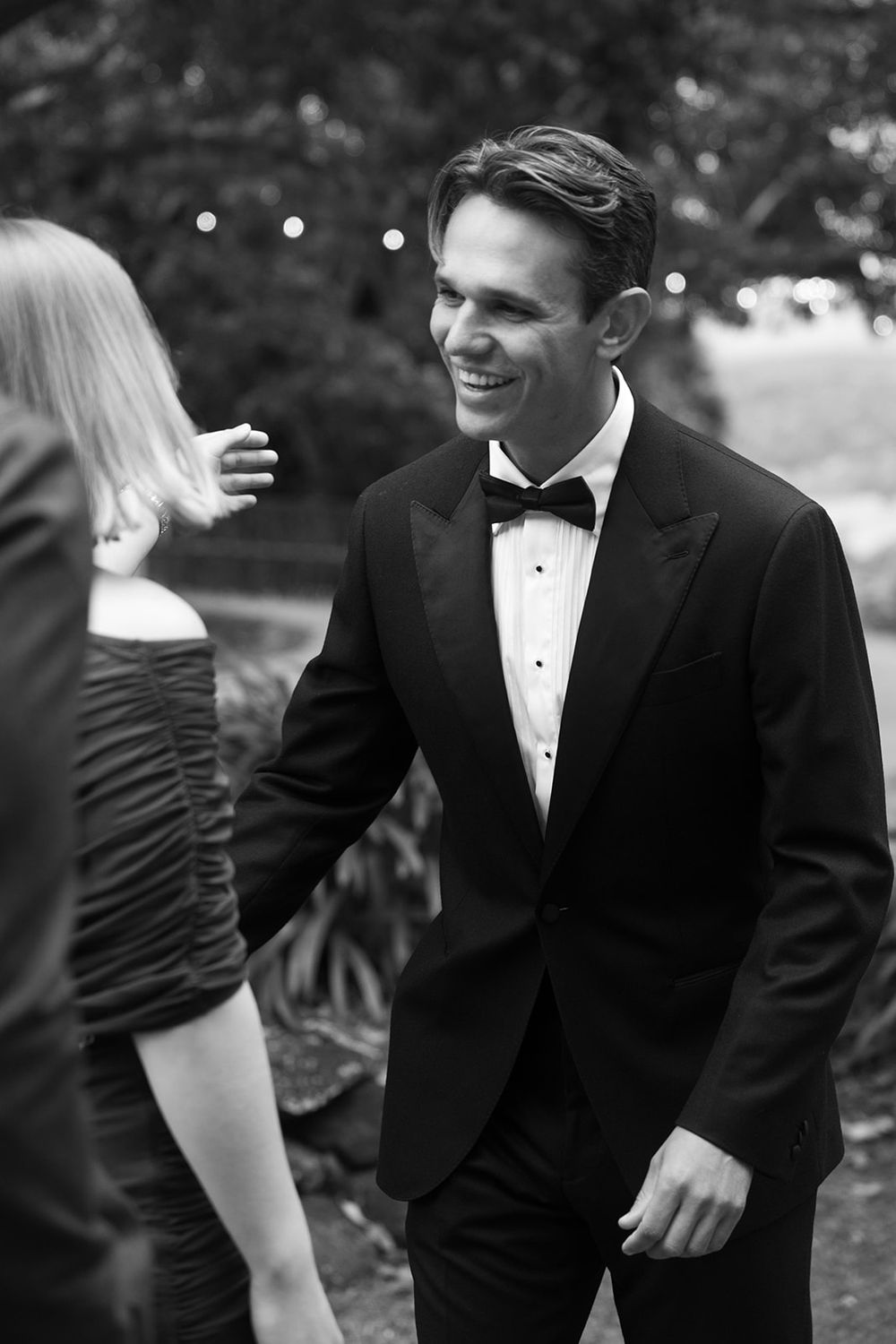 Smiling man in a black tuxedo and bow tie greets a woman in an off-the-shoulder dress at an outdoor evening event, black and white.