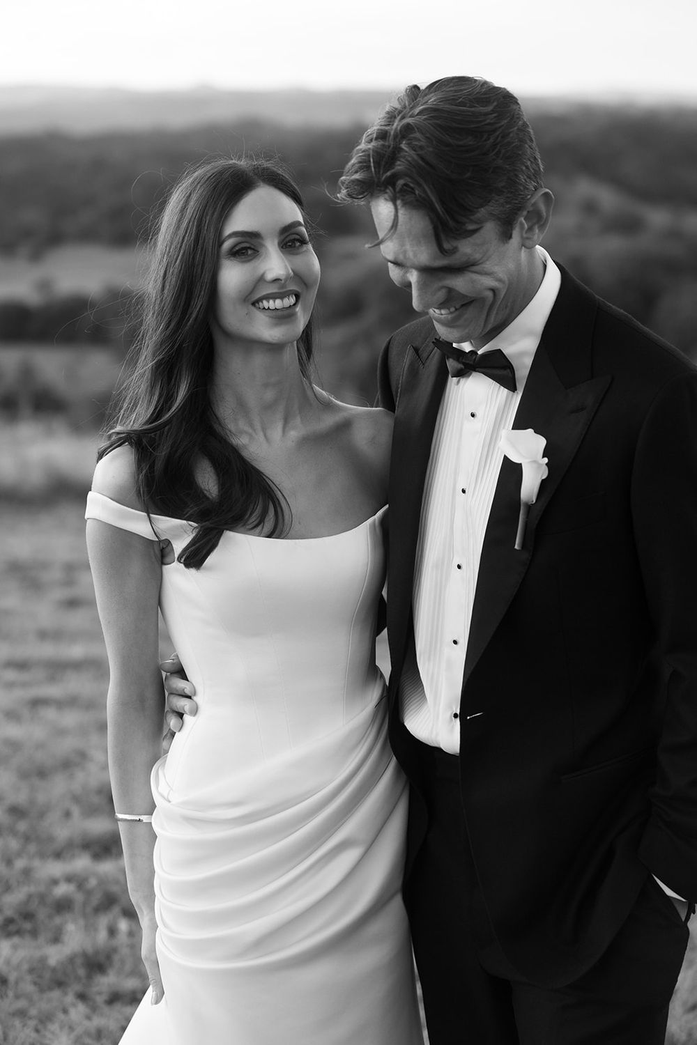 Black and white wedding portrait of a smiling bride in an off-shoulder satin gown embracing her groom in a tuxedo and bow tie outdoors.