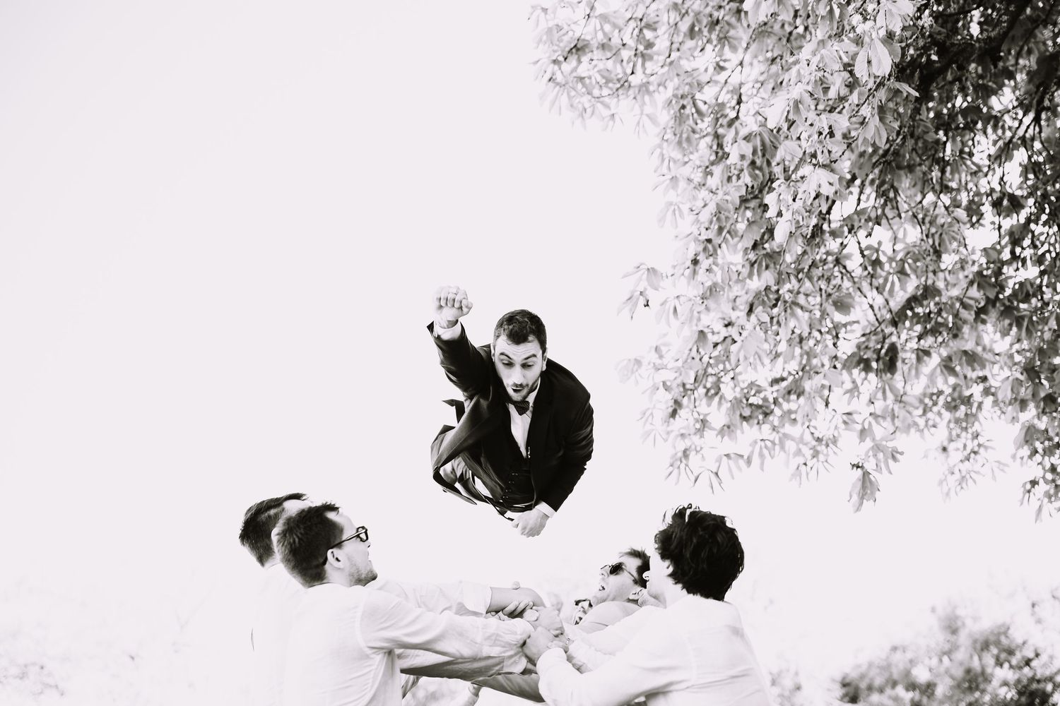 Photo noir et blanc du marié lancé en l'air par ses amis sous un arbre