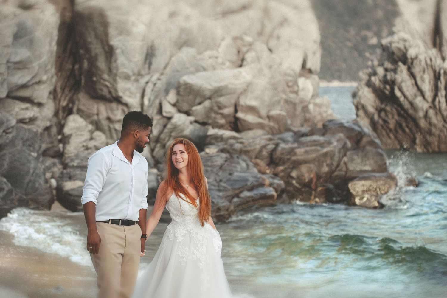Candid beach wedding photography of a bride and groom walking hand in hand on a rocky seashore.