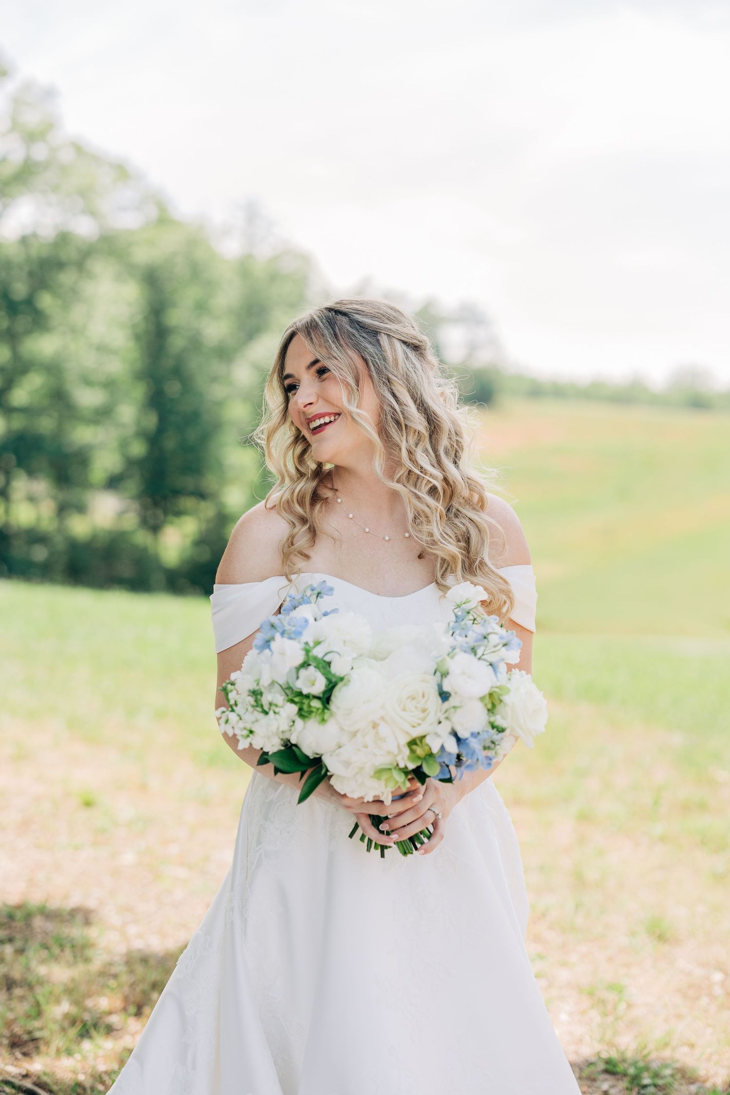 Smiling bride in off-shoulder white gown holds blue and white floral bouquet outdoors in a lush green field.