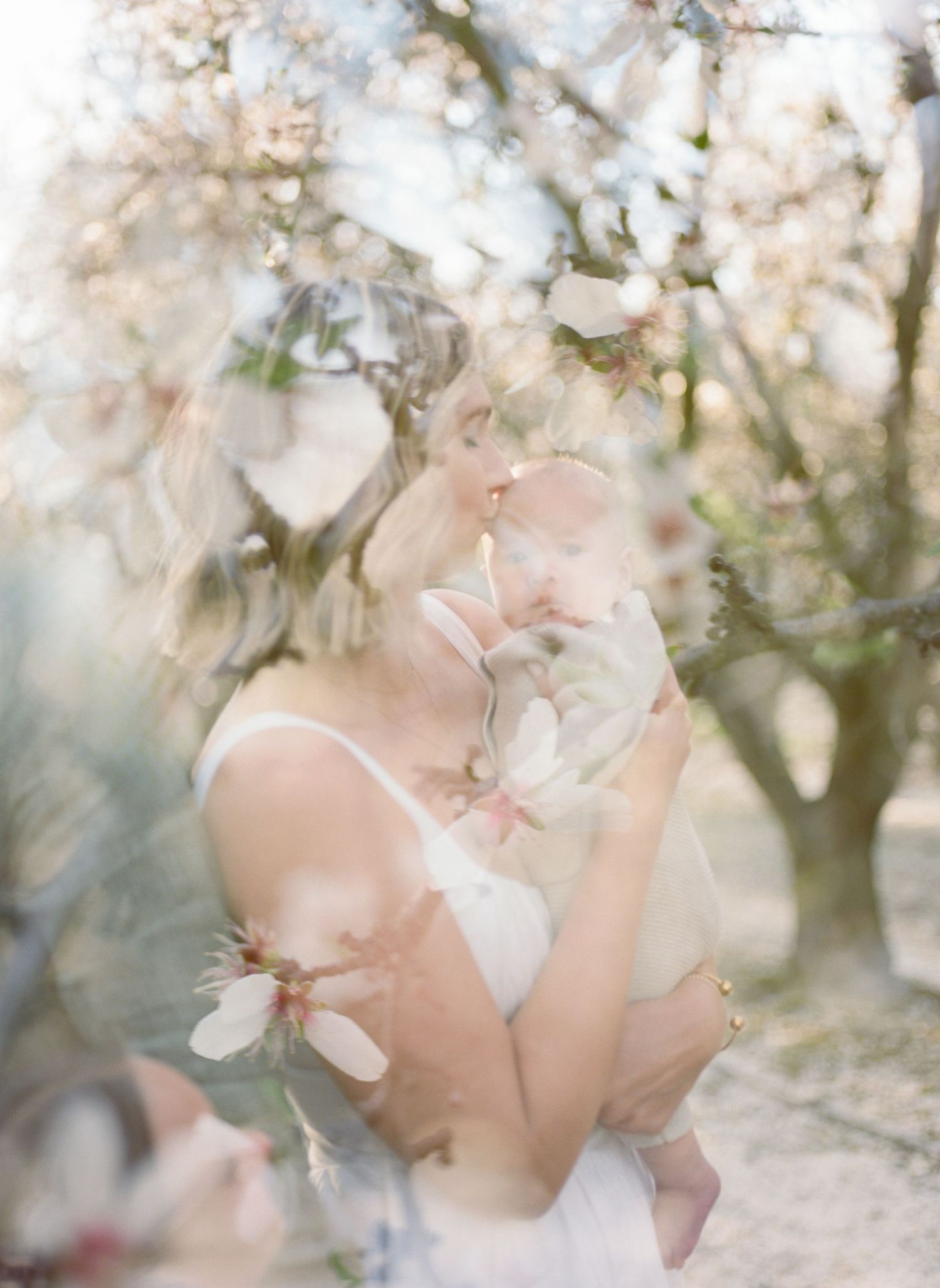 Mother kissing newborn baby among blooming almond blossom trees in a dreamy, soft-focus spring outdoor portrait in a double exposure frame