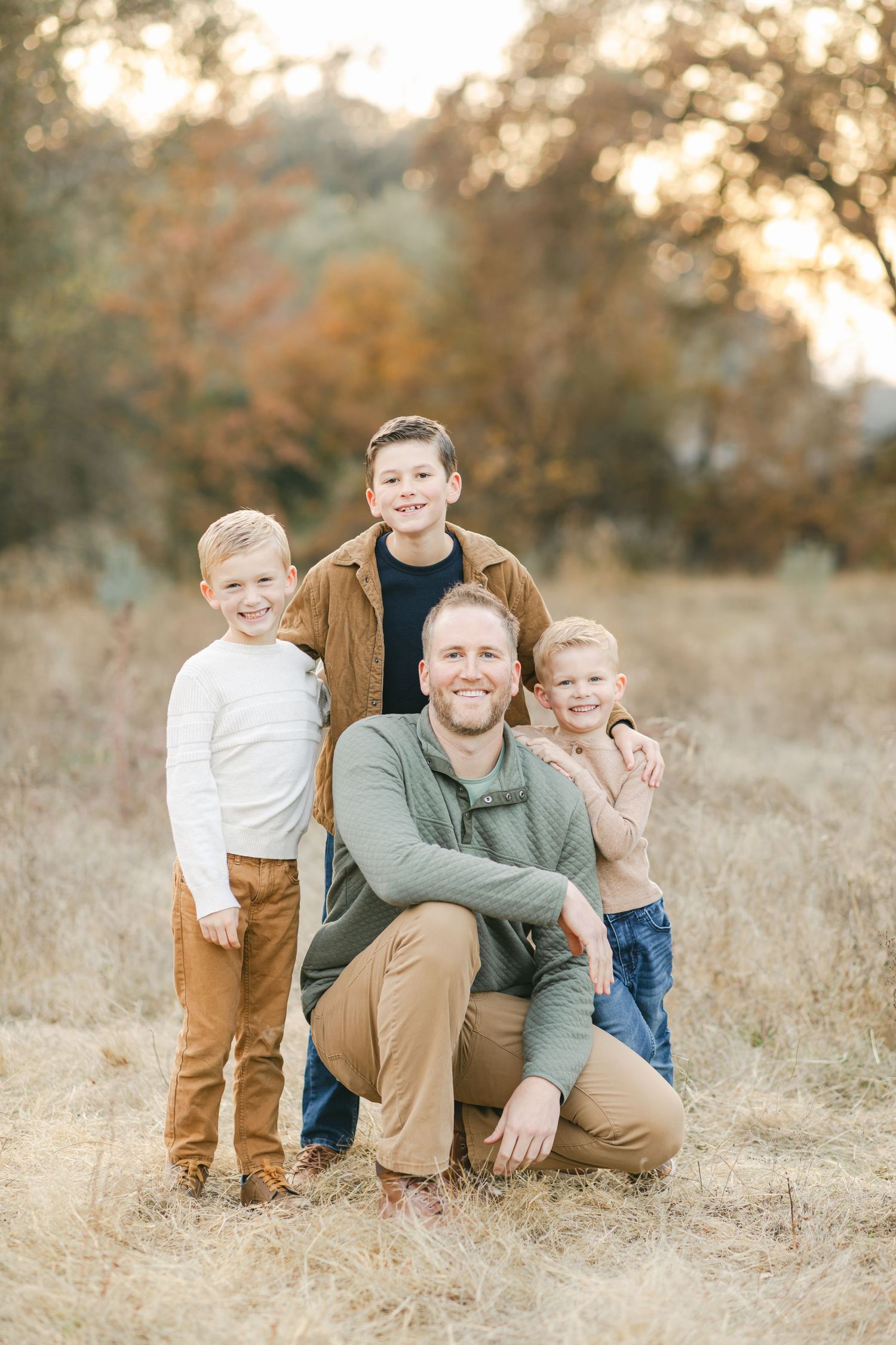 A father crouches in an autumn field smiling with his three young sons gathered around him.