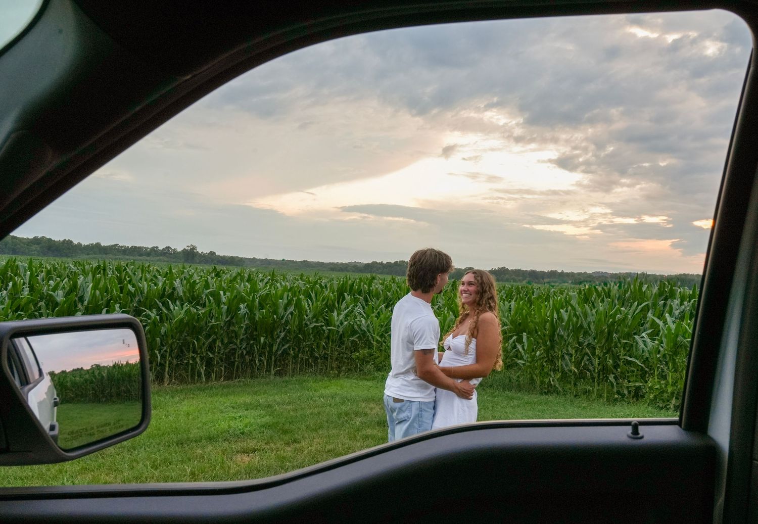 Couple embracing near a cornfield at sunset, viewed through a car window with mirror visible.