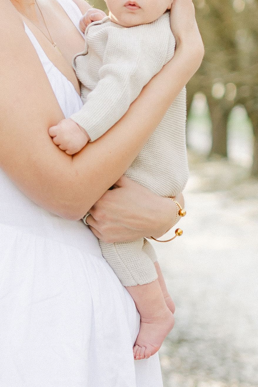 A mother in a white dress holds her newborn baby dressed in a cream knit onesie outdoors.
