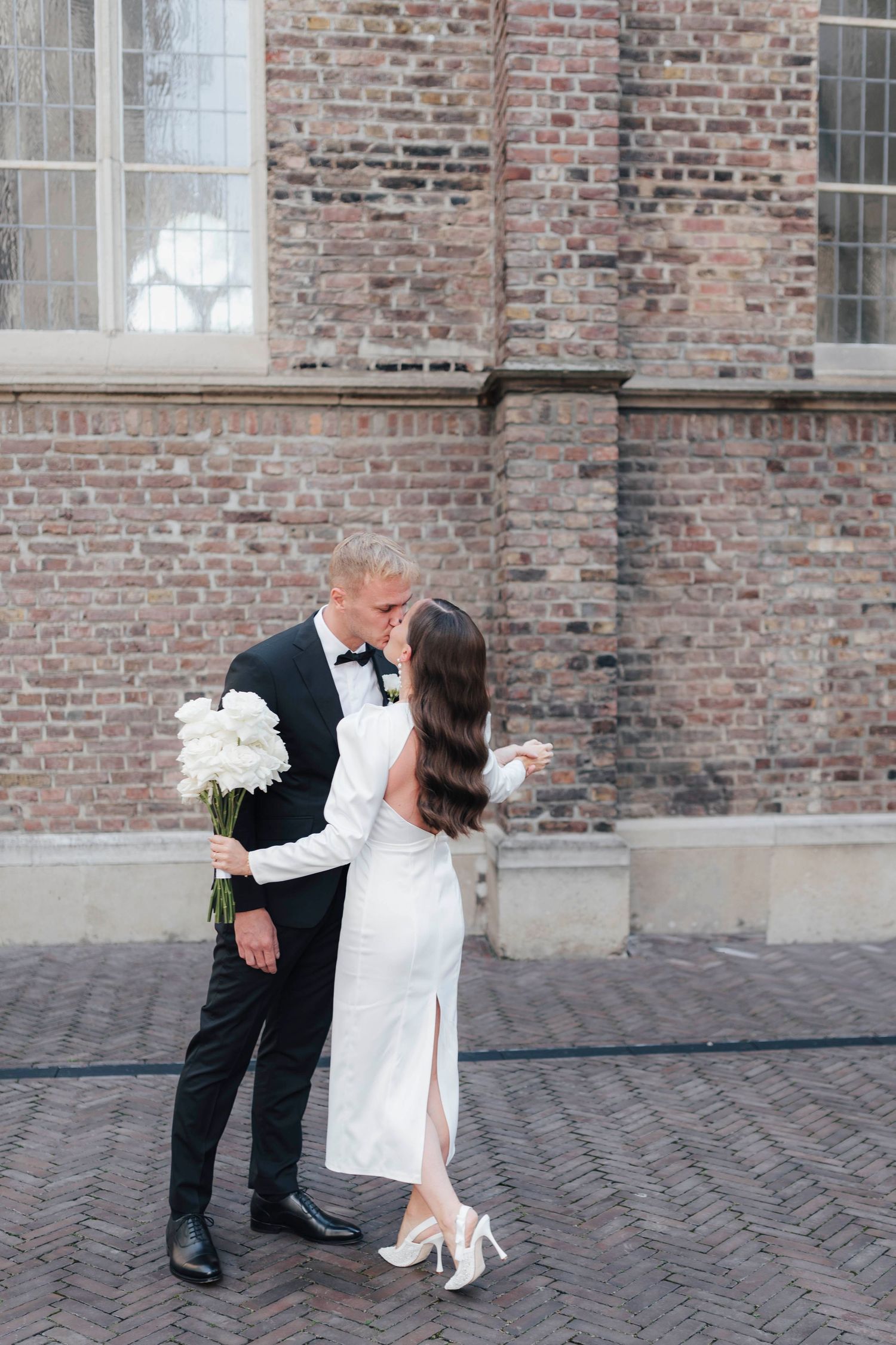 Bride and groom sharing a kiss outdoors near a brick building, bride holding white peonies bouquet in elegant white gown.
