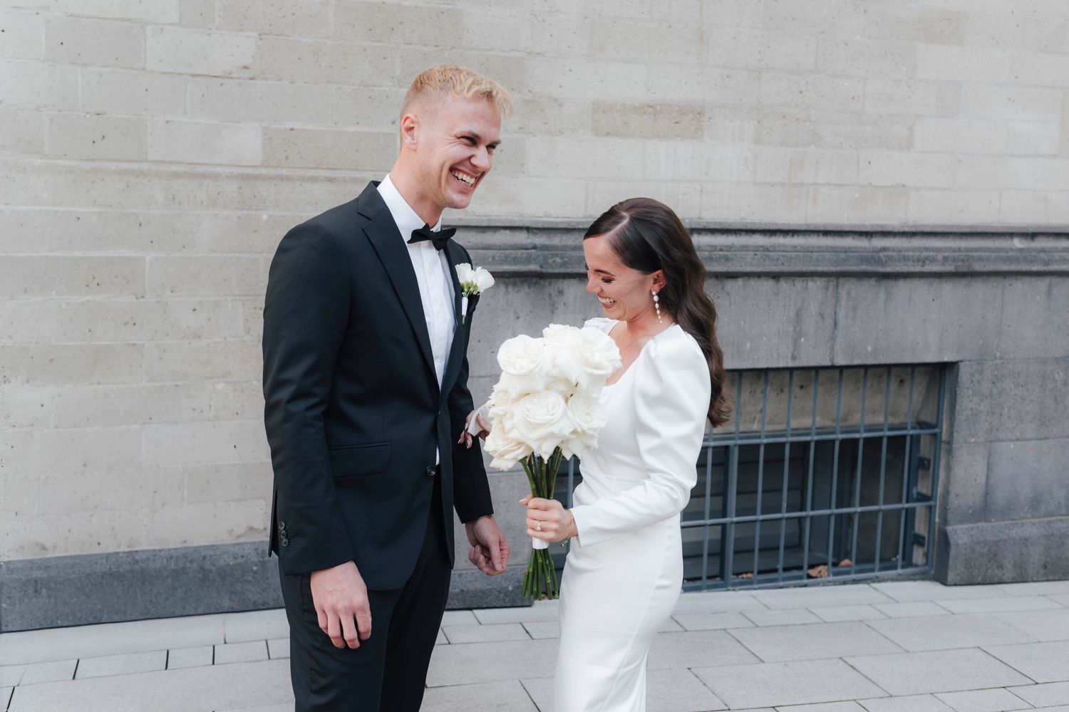 A smiling groom in a black tuxedo and bride in a white gown holding white peony bouquet stand together outdoors.