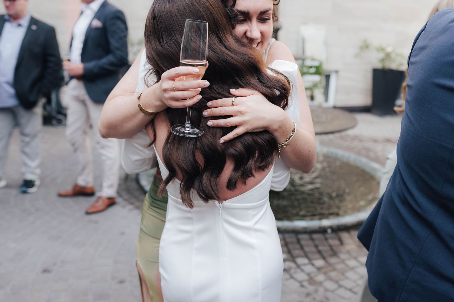 Two women share a warm hug at a wedding reception, one in a white dress holding a champagne flute.