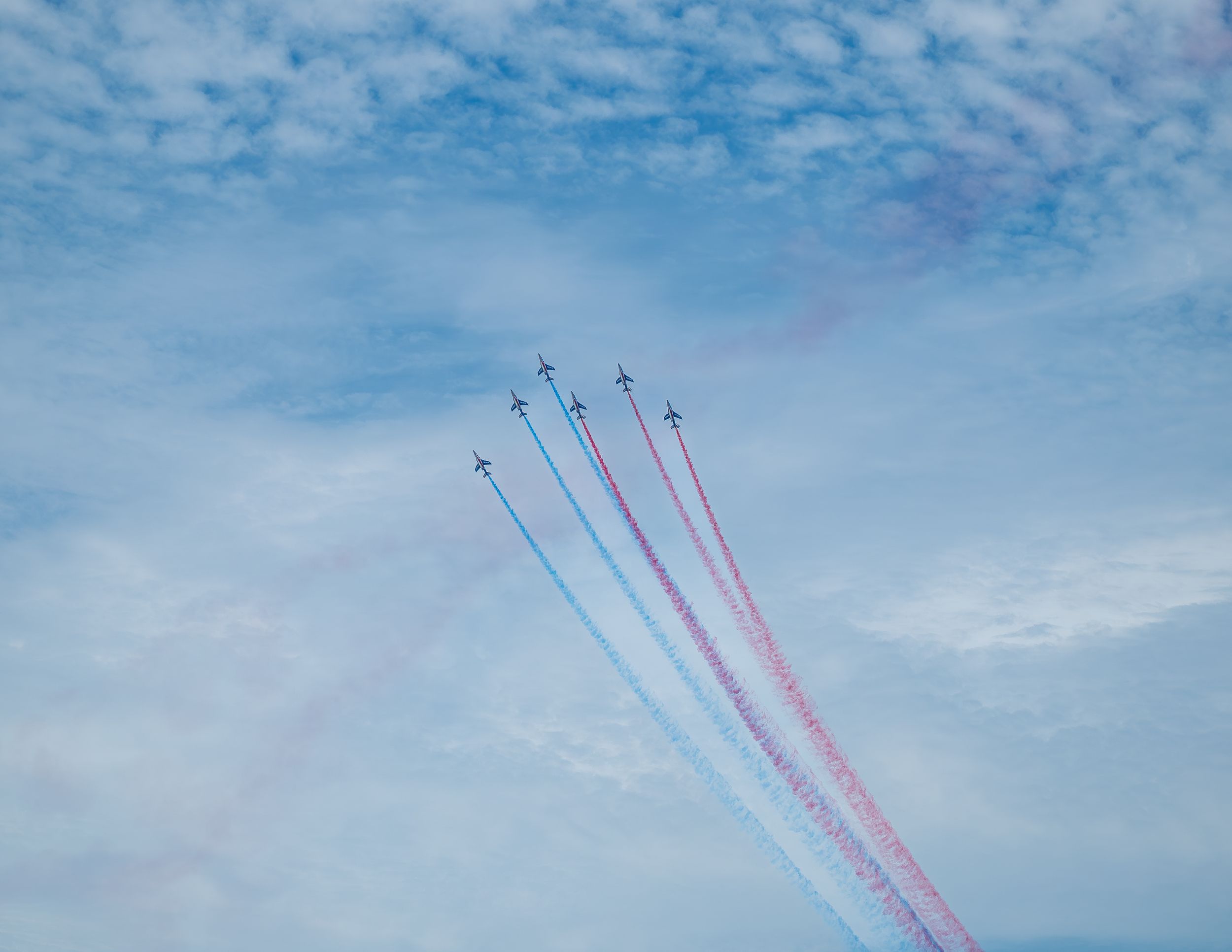 Five jets perform a formation flight leaving red, white, and blue smoke trails against a cloudy blue sky.