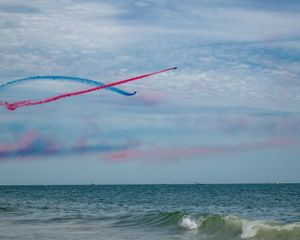 Aerobatic jets create colorful red and blue smoke trails over the ocean during a beachside airshow.
