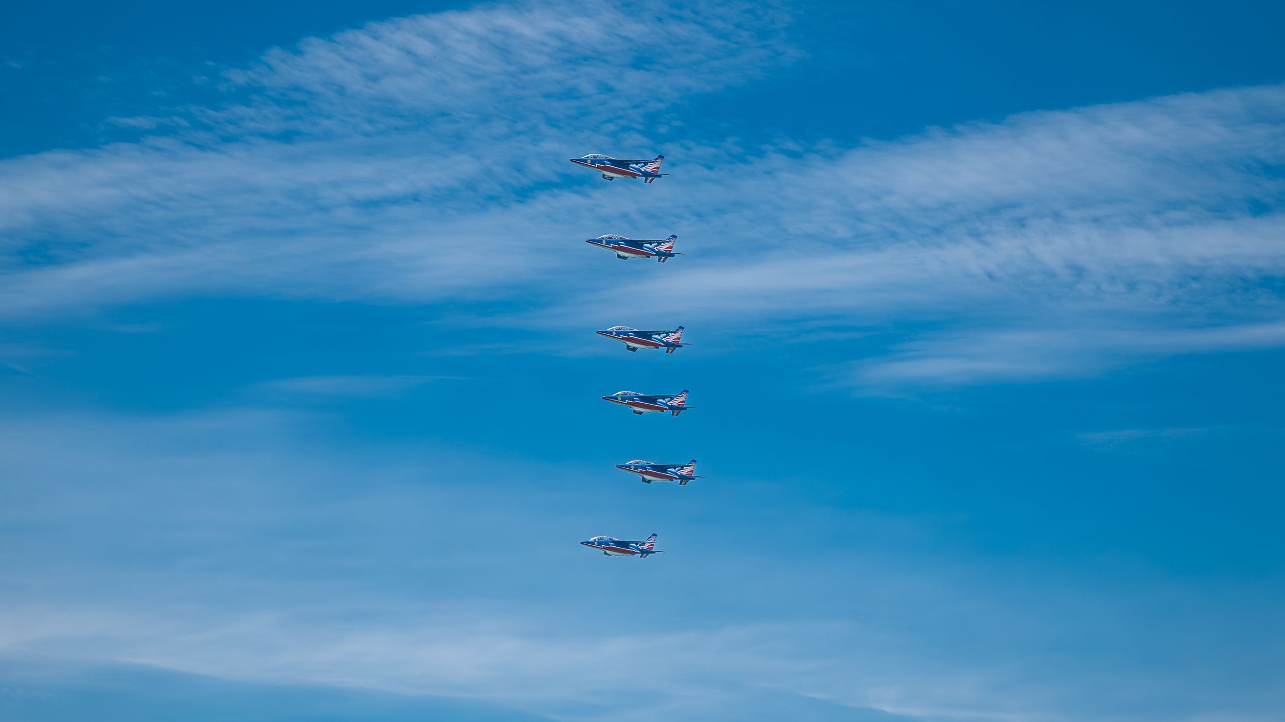 Six military jets fly in diagonal formation against a vivid blue sky with wispy clouds during an airshow.
