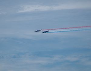 Four jets fly in formation at an airshow, trailing red, white, and blue smoke streams against a cloudy sky.