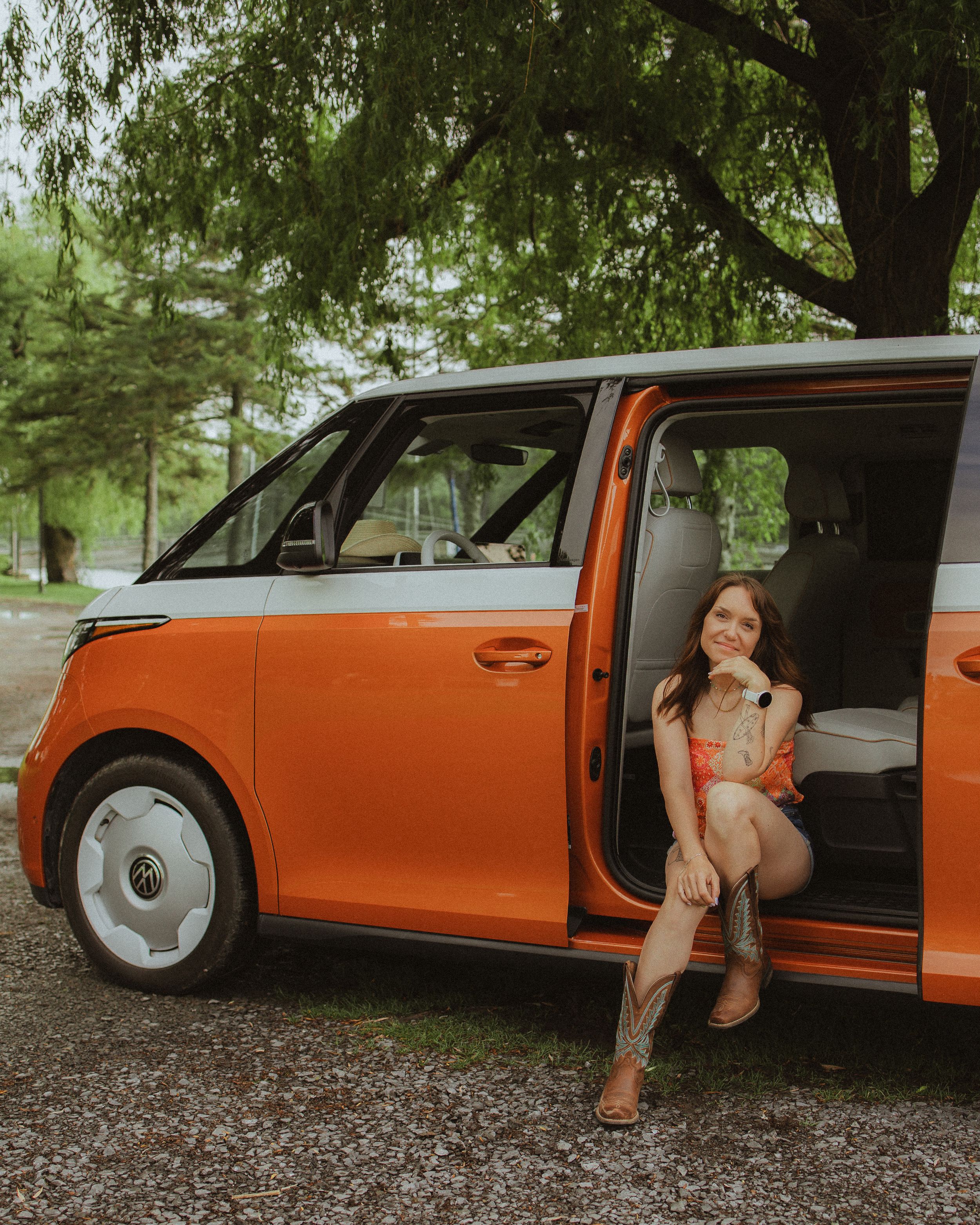 Smiling woman in cowboy boots sitting in open door of orange Volkswagen ID.Buzz van under green trees.