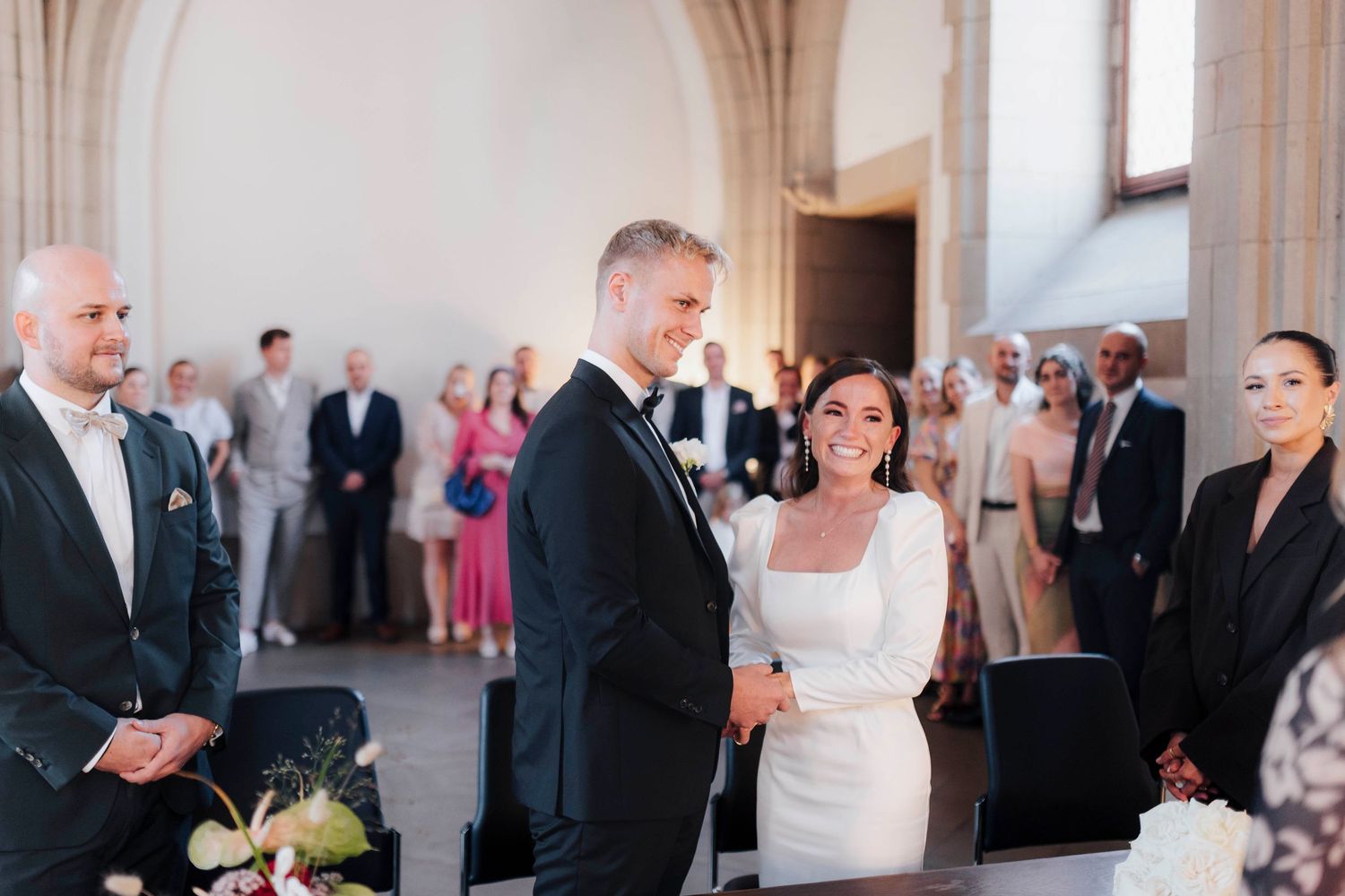 Couple smiling during elegant indoor wedding ceremony with guests standing in background in a bright venue.