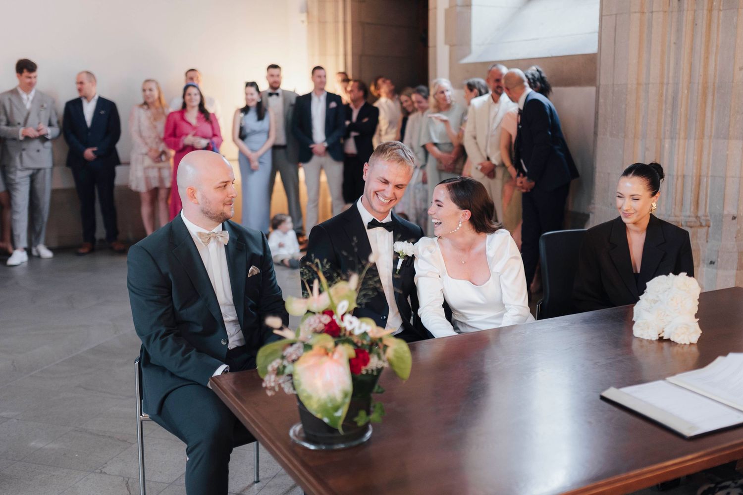 Bride and groom sit at signing table with guests standing behind during elegant indoor wedding ceremony.