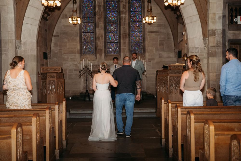 Bride walking down the aisle with escort in a stone church with stained glass windows during a wedding ceremony.