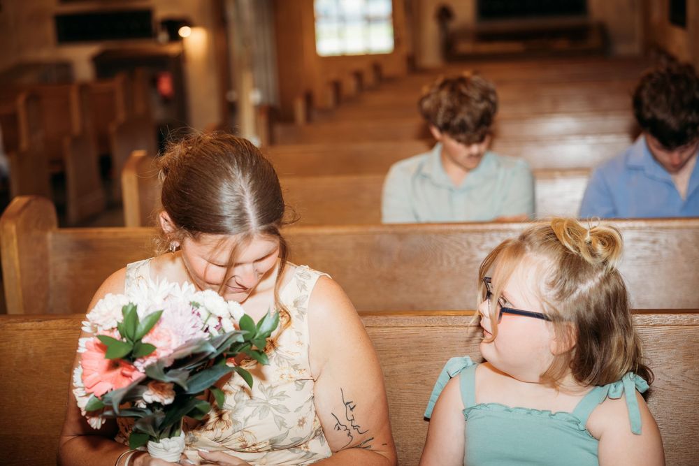 Woman holding bouquet smiles at young girl with glasses sitting together on wooden church pews at a wedding.