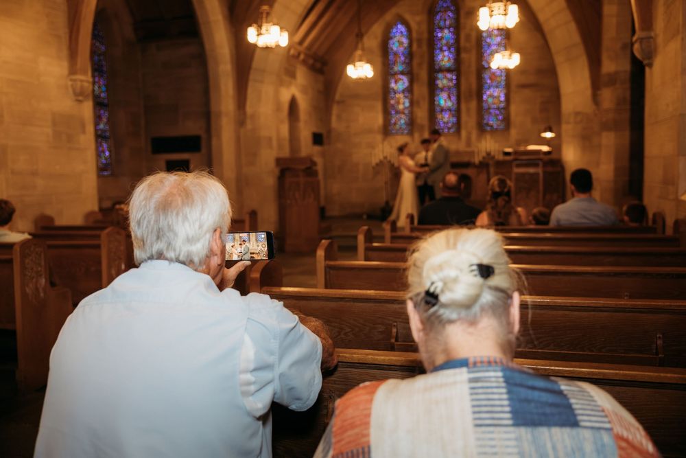 Elderly guests seated in church pews, one photographing a wedding ceremony with a smartphone near stained glass windows.