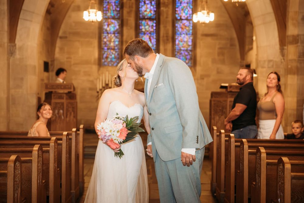 Bride and groom share first kiss at church altar, bride holding pink bouquet, stained glass windows in background.