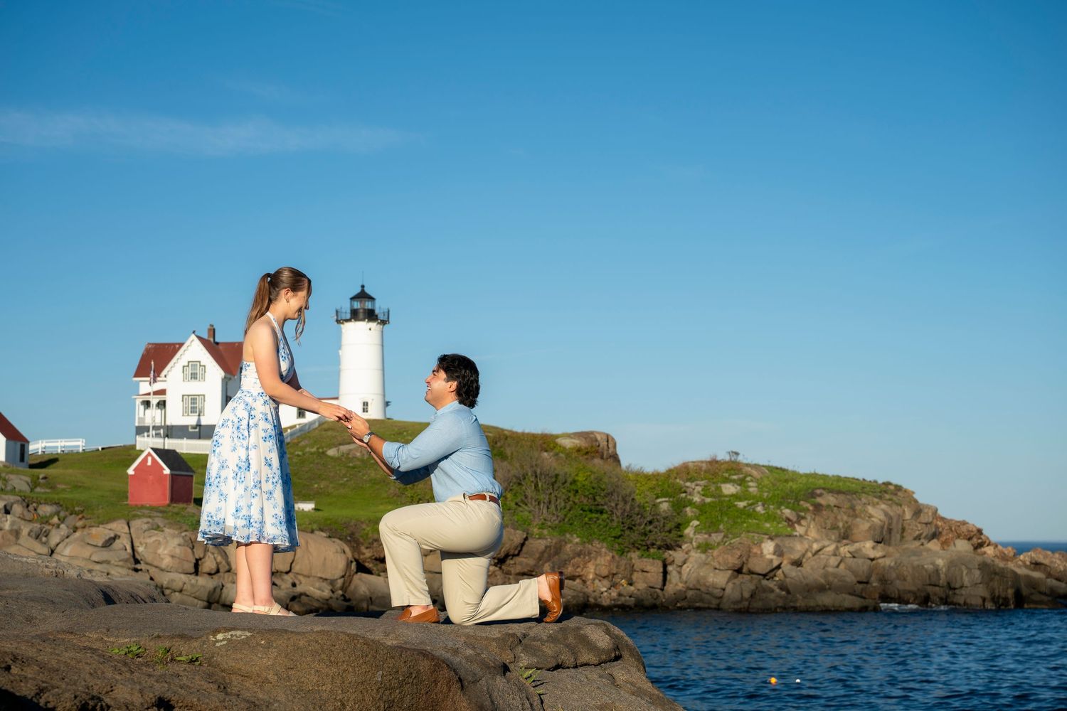 Man proposing to woman on rocky shore with lighthouse backdrop on a sunny day.
