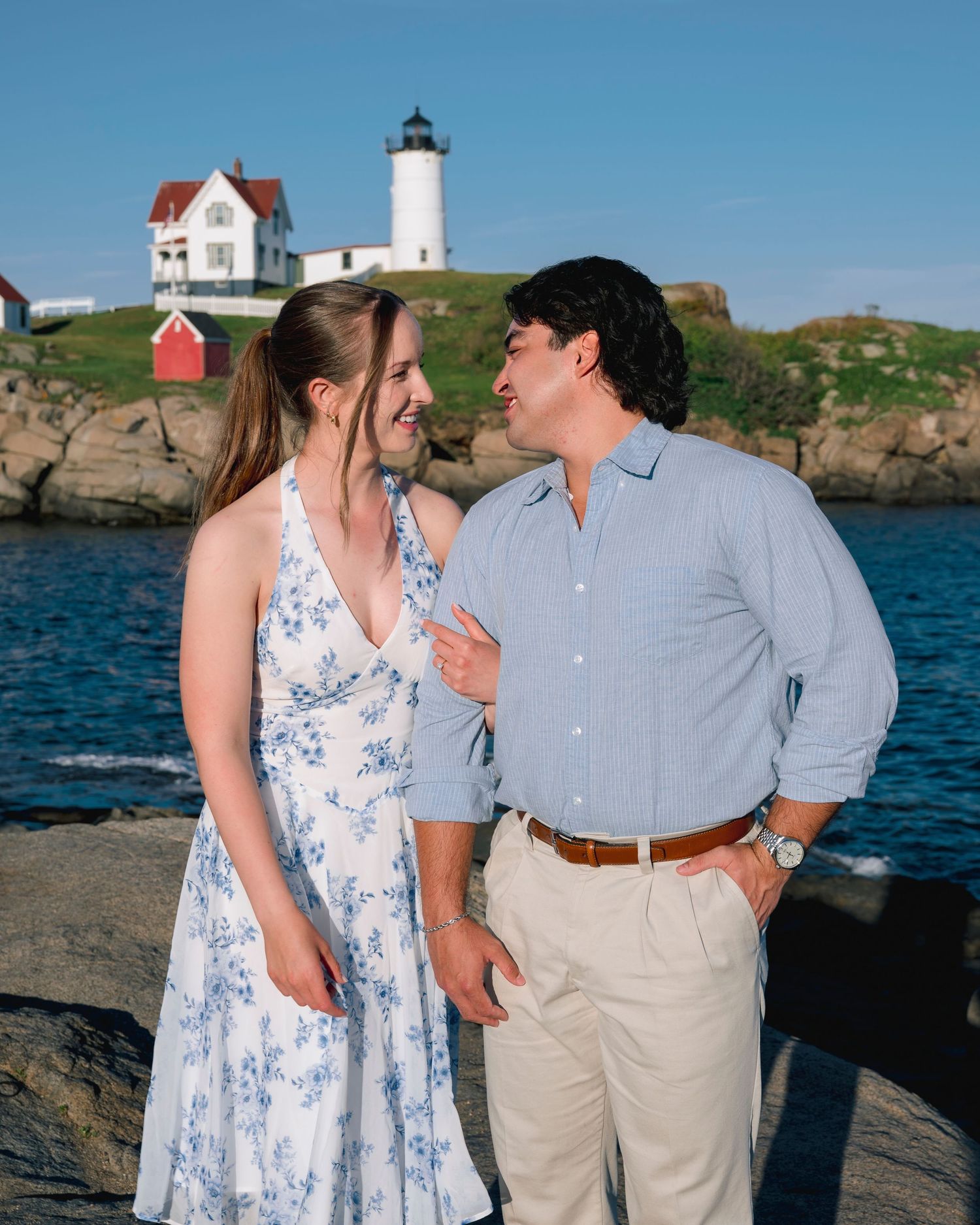 Couple smiling at each other near rocky New England coastline with historic lighthouse and house in background on sunny day.
