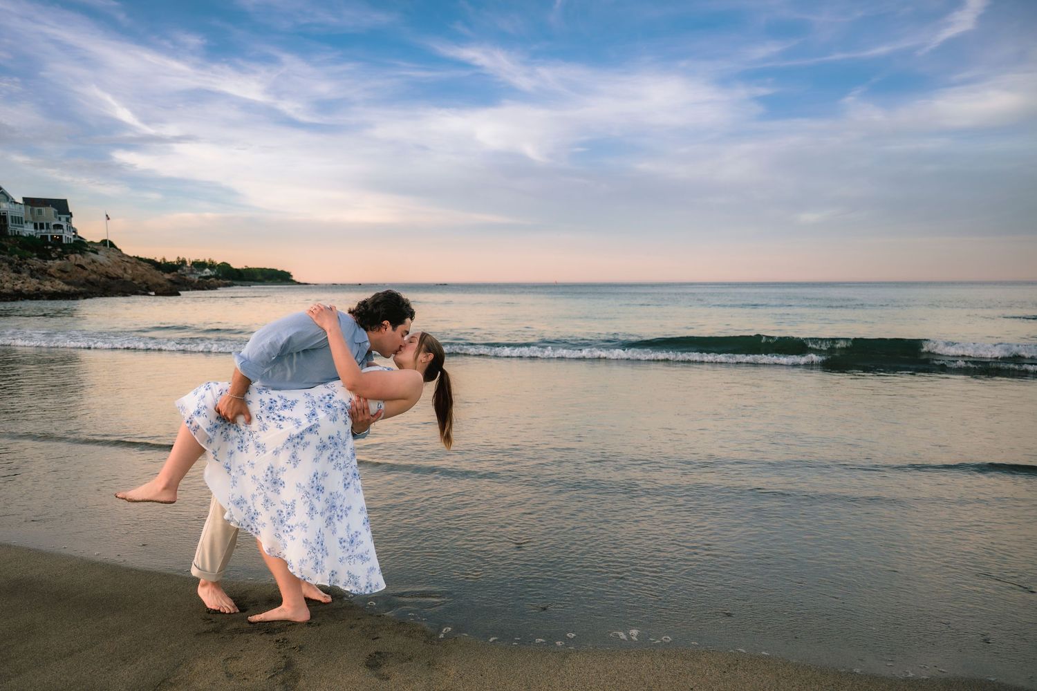Couple sharing a romantic dip kiss on a sandy beach at sunset with ocean waves and cloudy sky behind them.