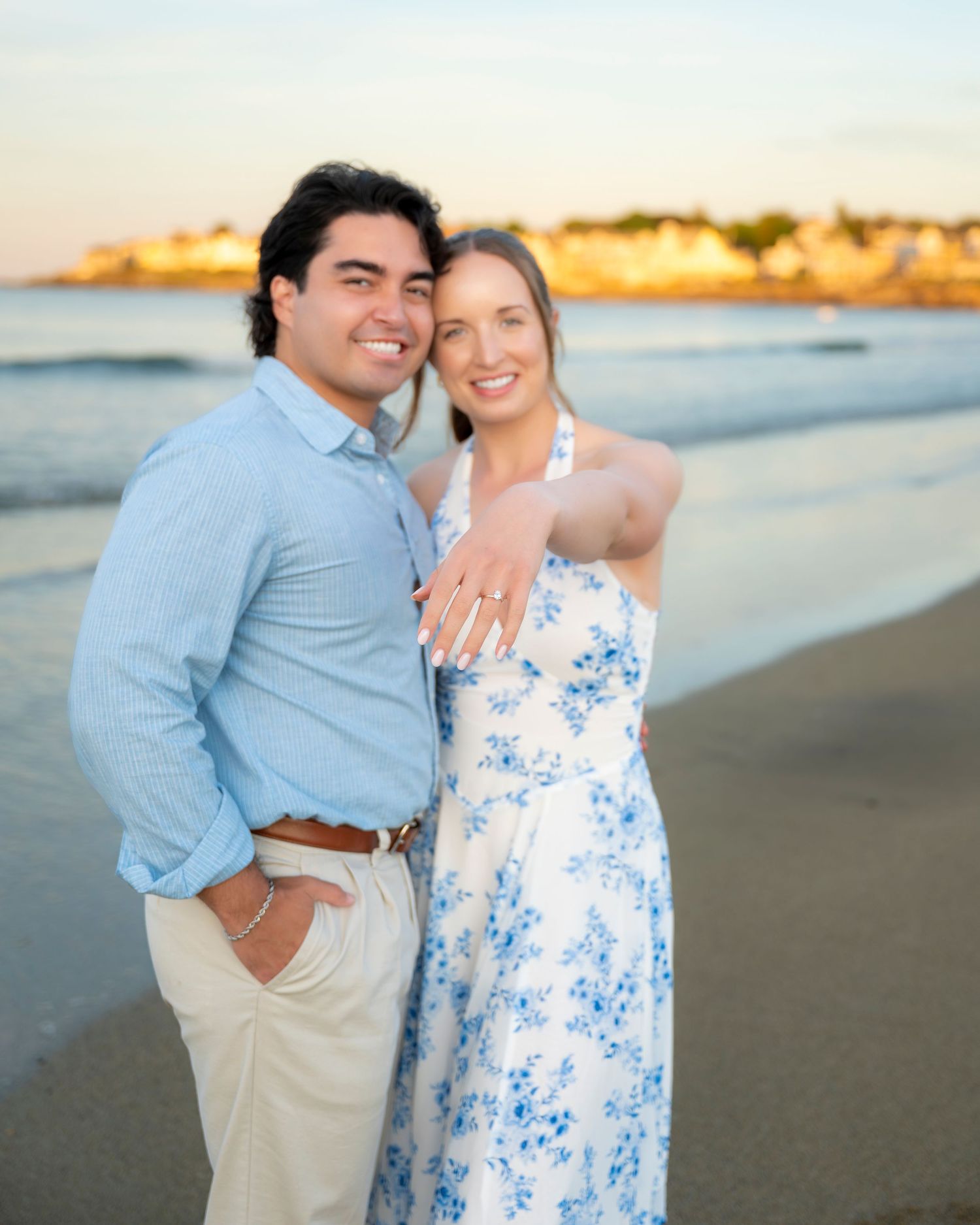 Happy couple embracing on a sandy beach at sunset, woman wearing a white floral dress showing engagement ring.