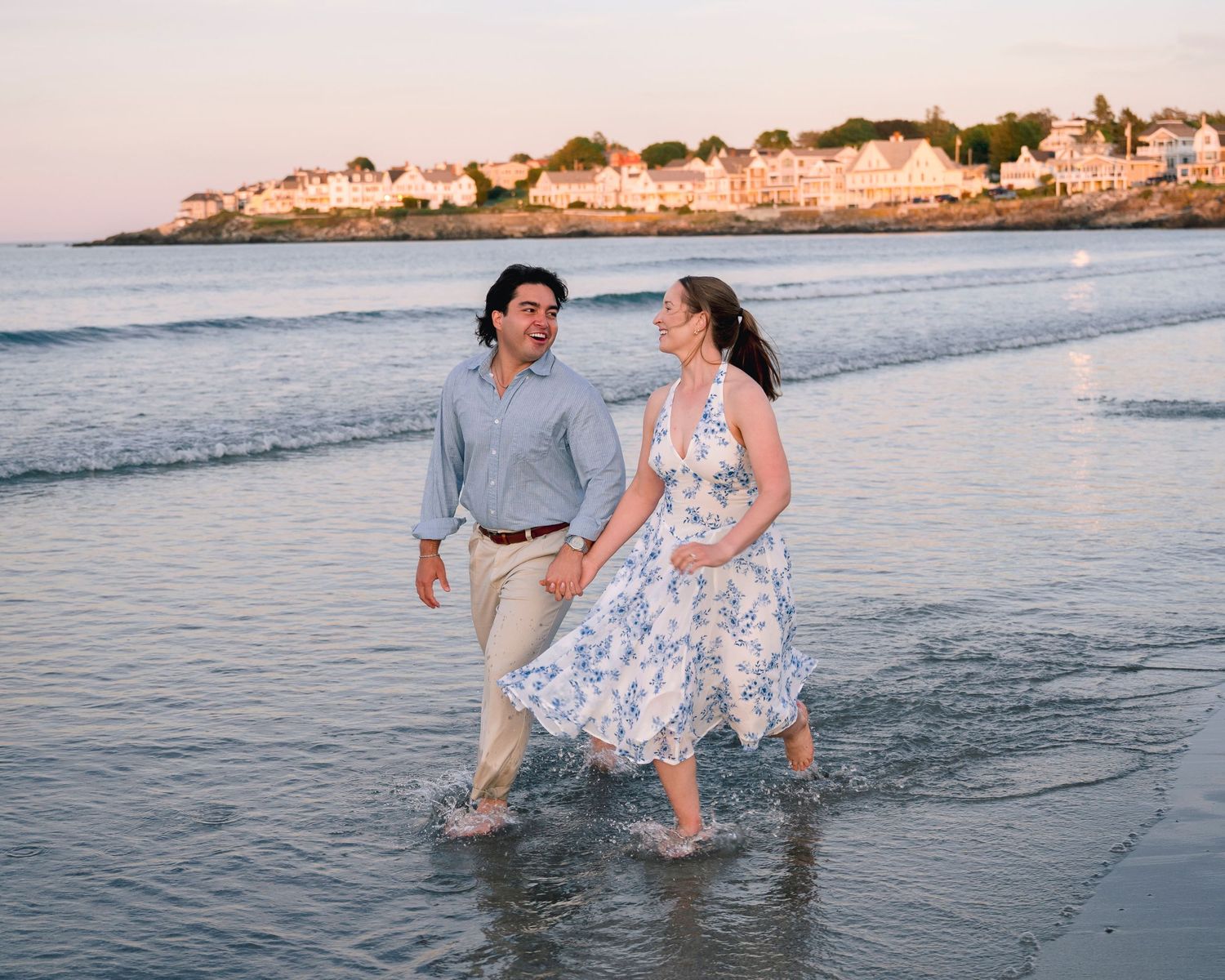 Couple walking hand-in-hand on a sandy beach at sunset, woman in floral dress, coastal town in background.