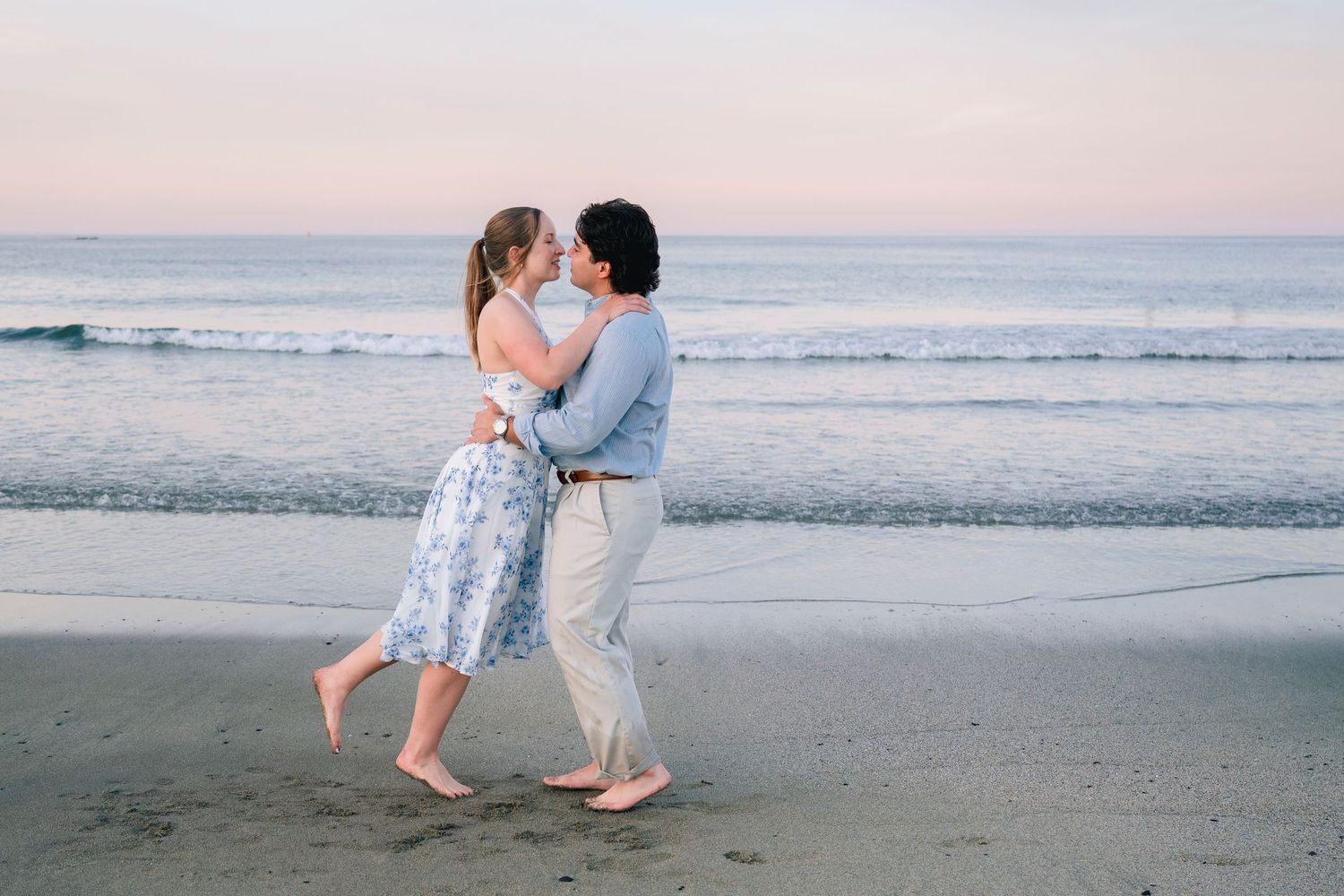 Couple dancing barefoot on a sandy beach at sunset, embracing romantically with ocean waves in the background.