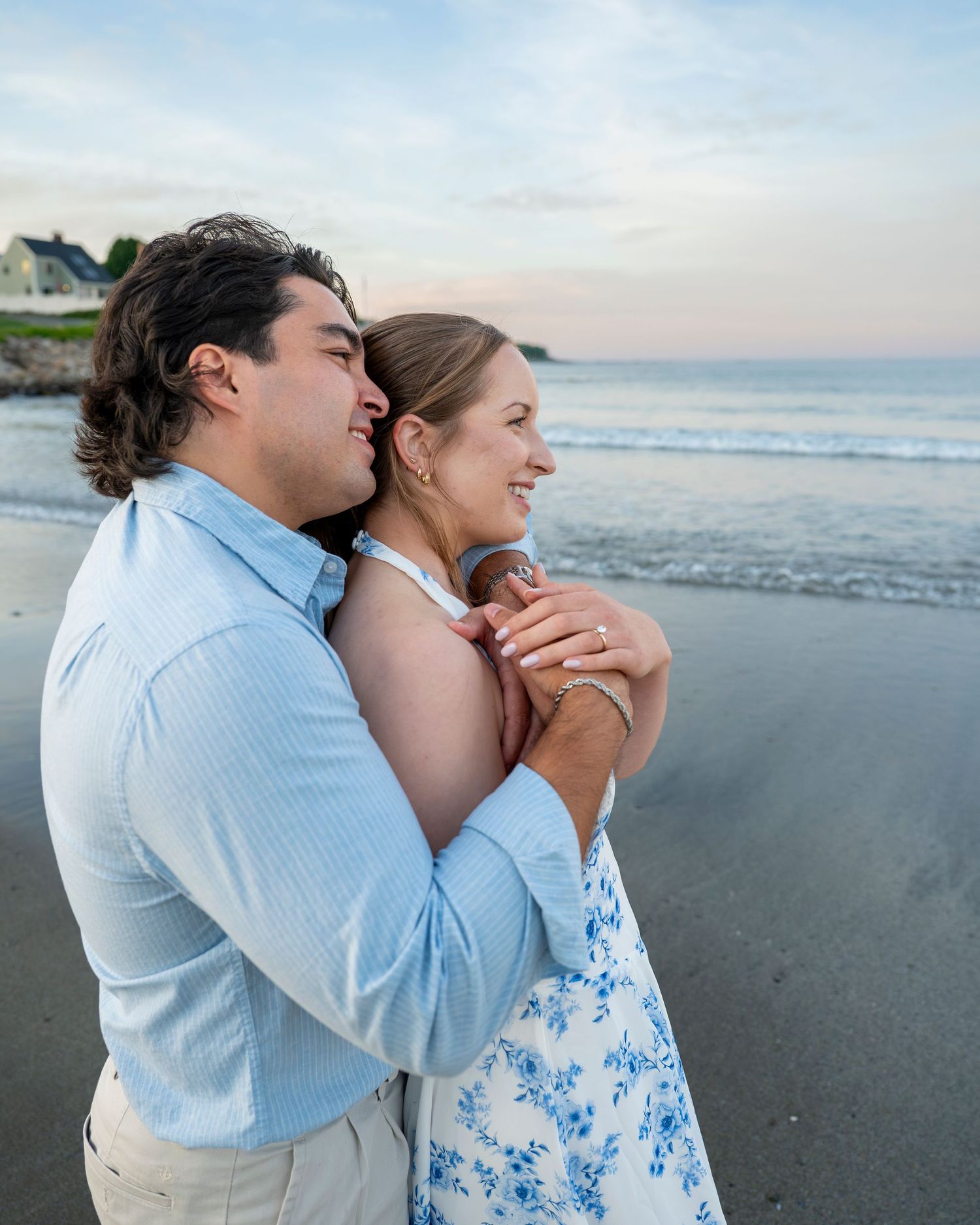 Couple embracing on a beach at sunset, woman in floral dress, man in light blue shirt, ocean waves in background.