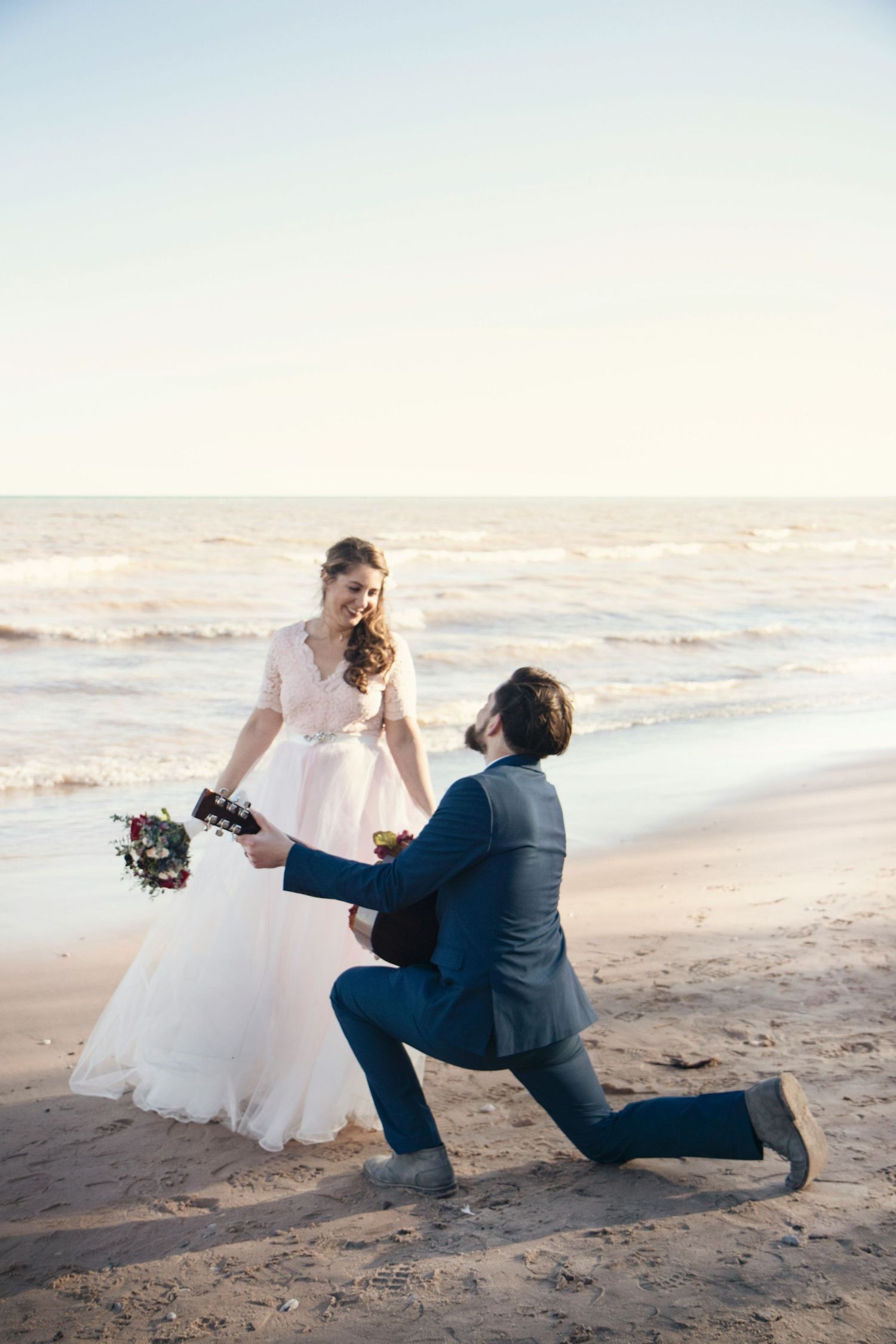 Groom kneeling on one knee before bride in white dress on sandy beach, ocean waves in background, romantic wedding photo.