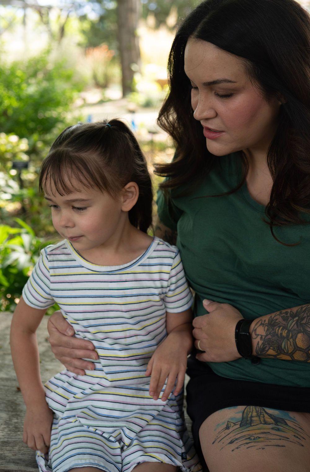 A woman in a green top with tattoos sits outdoors with a young girl in a striped dress.