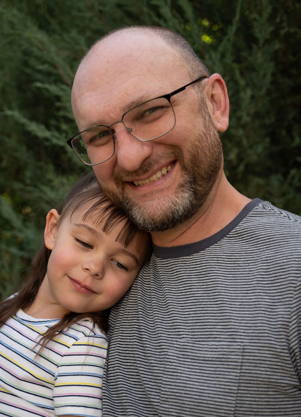 A bald man with glasses and a beard smiles warmly while a young girl leans her head against him outdoors.