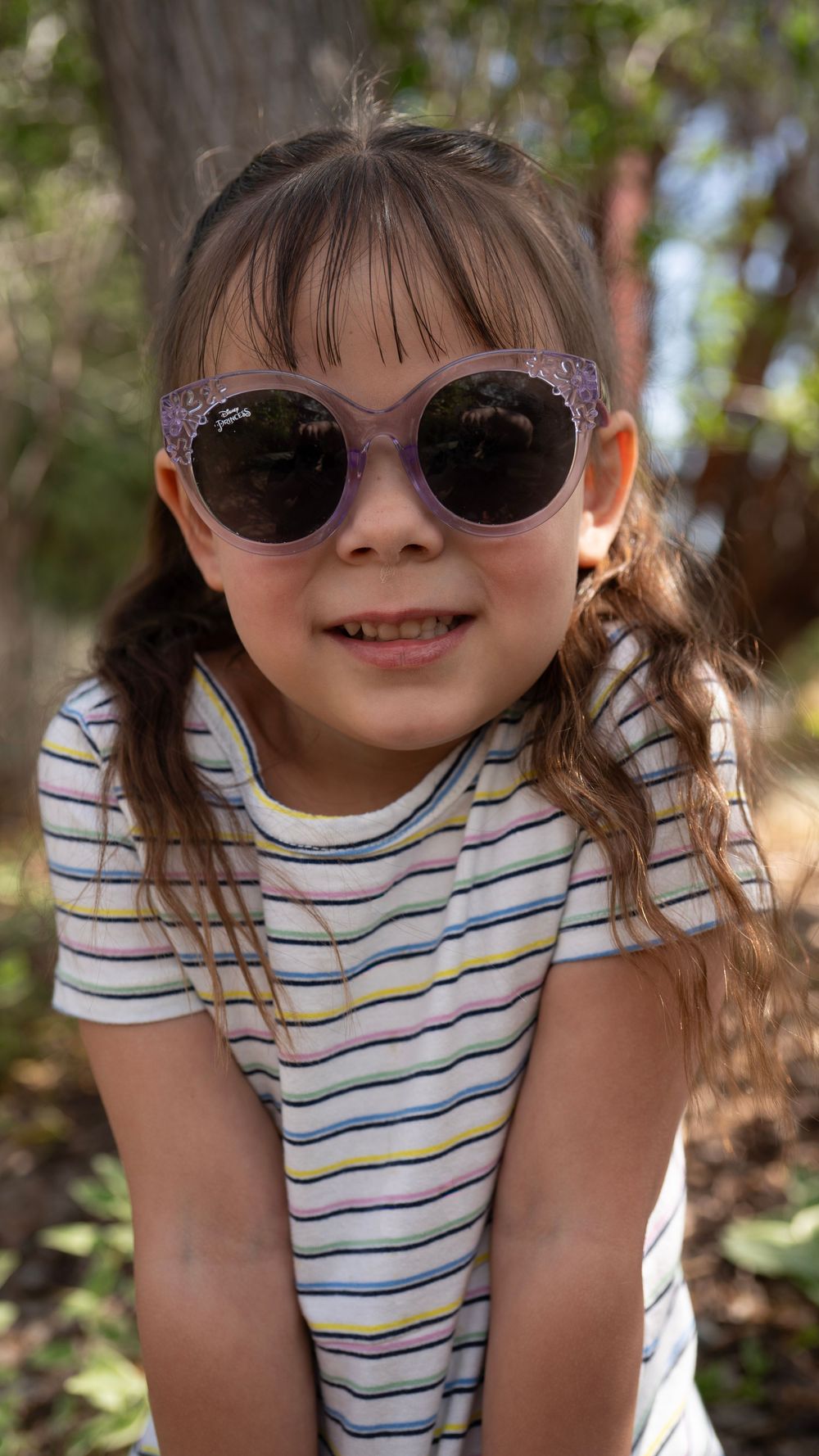Young girl smiling in purple cat-eye sunglasses and striped shirt outdoors among trees.