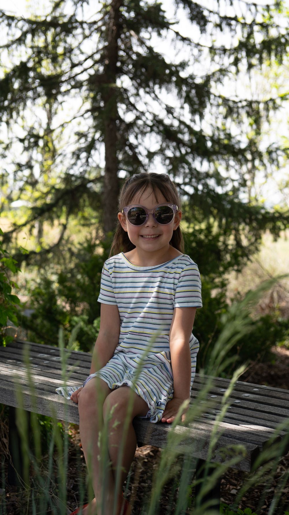 A smiling young girl wearing sunglasses and a striped dress sits on a wooden bench surrounded by lush green trees.