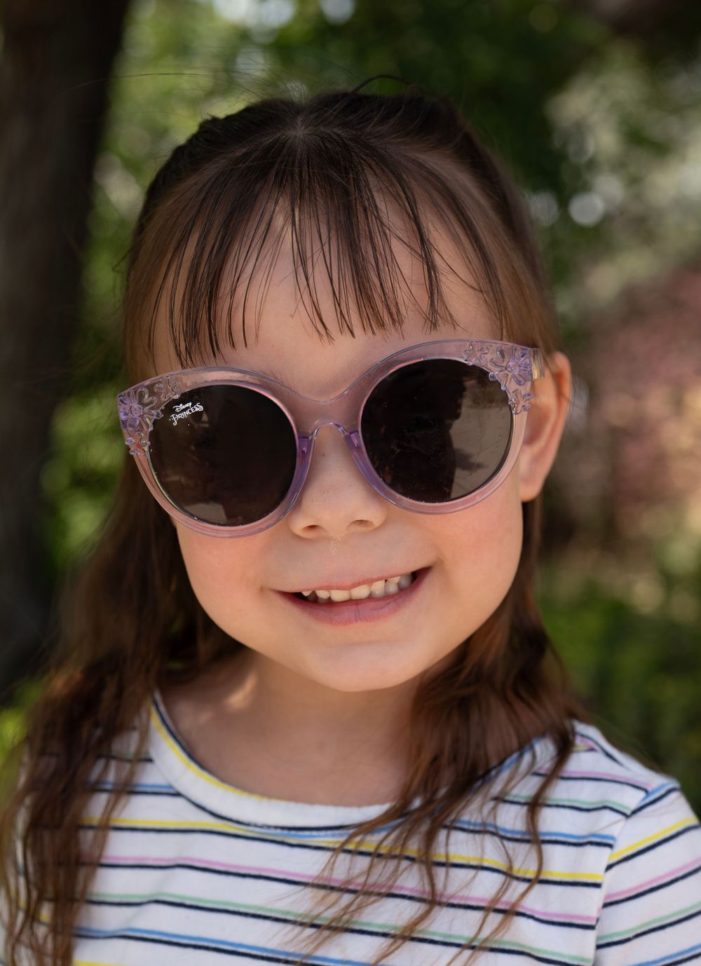A smiling young girl wearing purple glitter cat-eye sunglasses and a striped shirt outdoors.