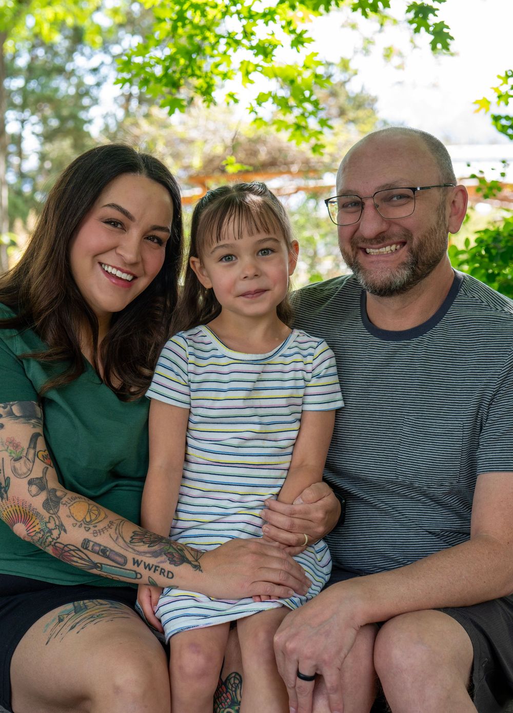 A smiling family of three poses outdoors, with a tattooed mother, young daughter, and bespectacled father seated together.