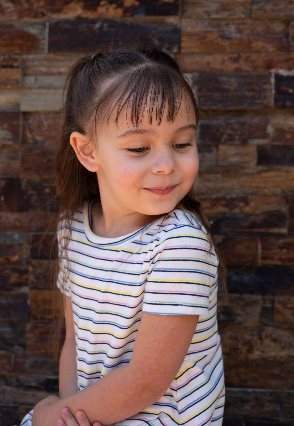 Young girl with bangs in striped dress smiling downward against stone wall backdrop.