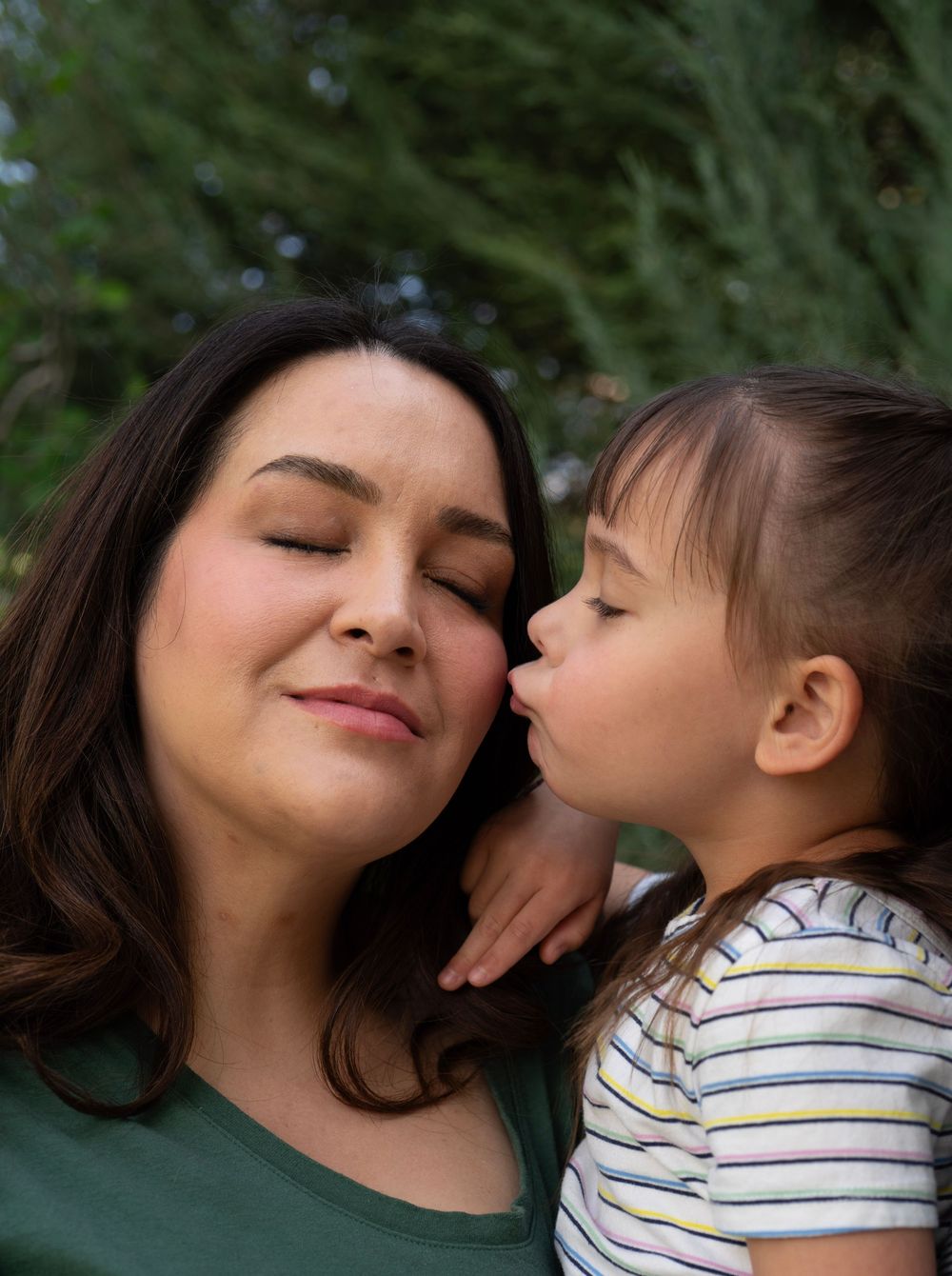 A young child tenderly kisses the cheek of a smiling dark-haired woman outdoors near green trees.
