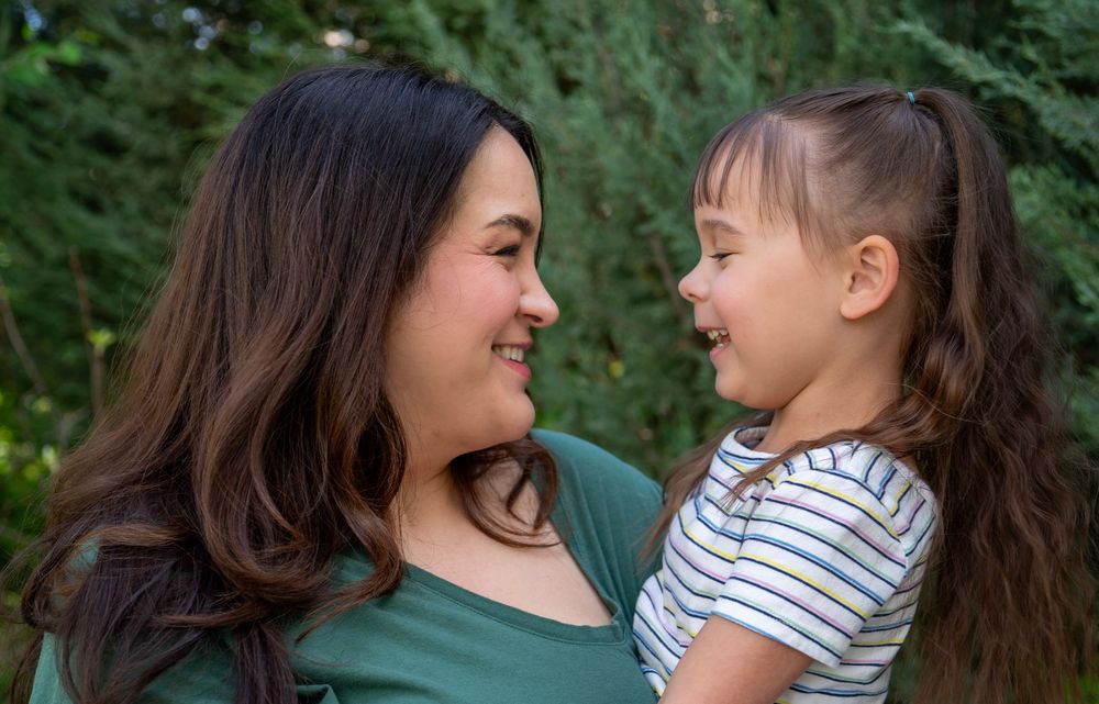 A smiling woman holds a happy young girl outdoors, sharing a joyful moment surrounded by green foliage.
