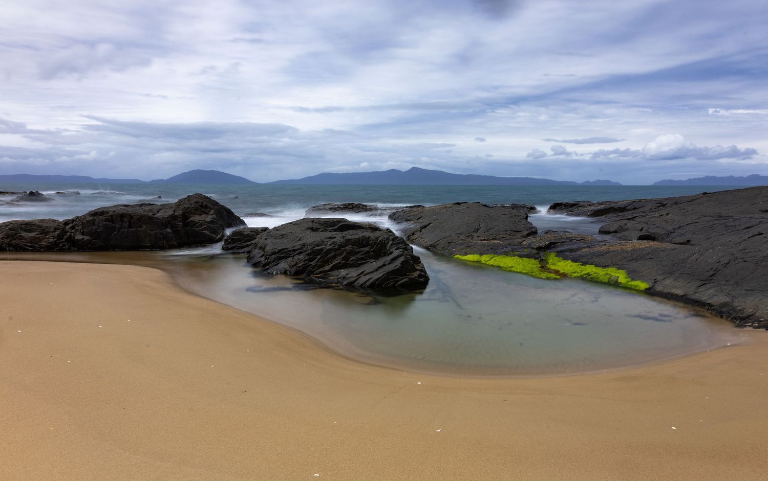 Rocky beach with green algae pools, sandy shore, and misty mountains under cloudy sky.