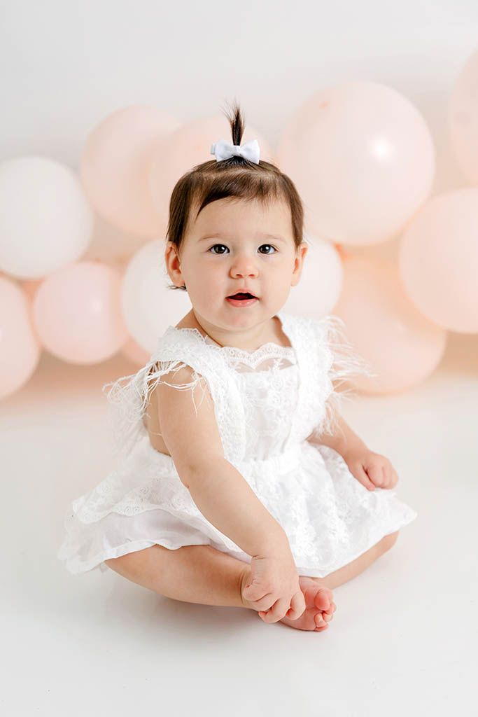 Baby girl in white lace dress sits on floor surrounded by pink and white balloons during first birthday photo session.